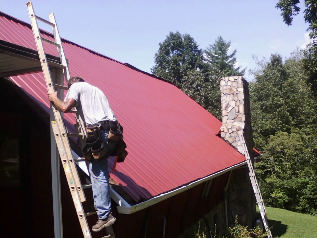 Roofer on a ladder beside a red metal roof and stone chimney — Soffit & Fascia Repair by Roofing Brothers in Richmond Dale